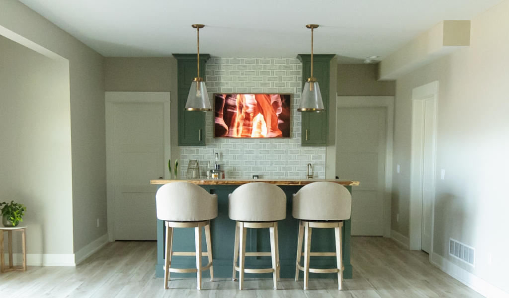 Modern home kitchen showcases a Byron Center Whole Home Remodel with three upholstered barstools at an island, pendant lights overhead, and a wall-mounted TV above a tiled backsplash.