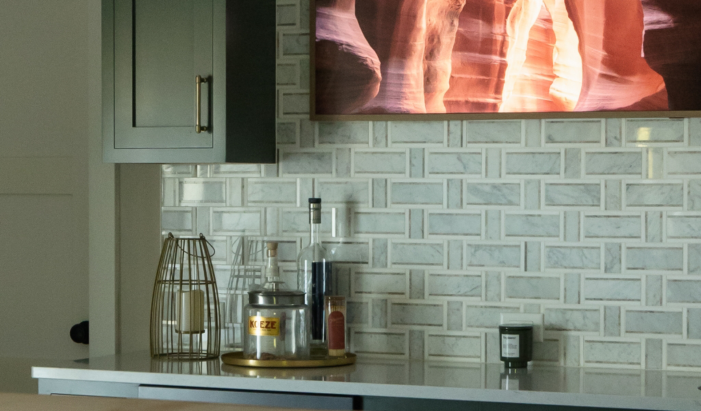 A kitchen counter from a Byron Center Whole Home Remodel features bottles, a candle, a wire utensil holder, and a tray, all set against a white tile backsplash with a framed picture above.