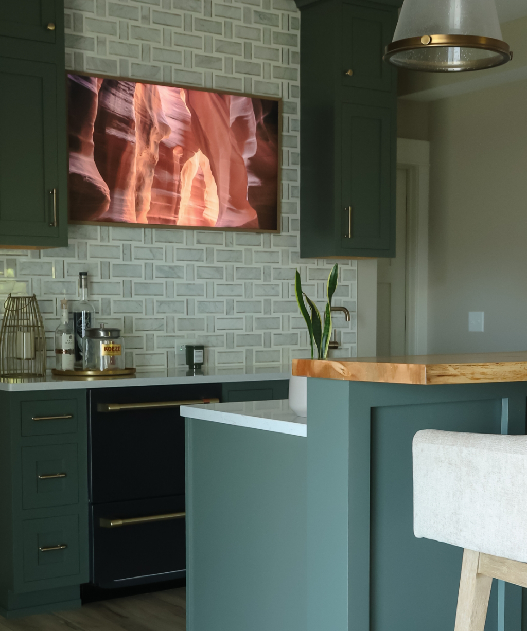 Modern kitchen from a Byron Center Whole Home Remodel featuring green cabinets, a patterned tile backsplash, wooden island, potted plant, and a wall-mounted TV displaying a canyon image.