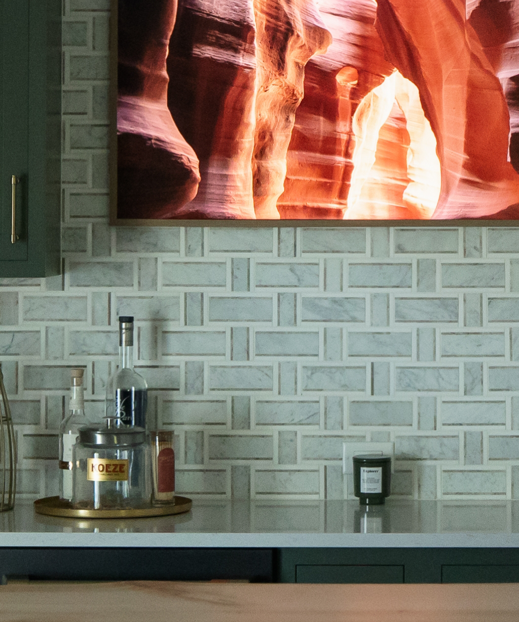 A kitchen countertop with bottles and a candle, backed by a white geometric tile backsplash and a framed photo of a canyon, showcases the refined style of this Byron Center Whole Home Remodel.