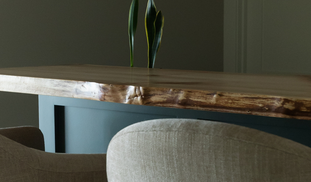 A close-up of a wooden table with a natural edge, two beige upholstered chairs, and a green plant in the background adds inviting style to this Byron Center Whole Home Remodel.
