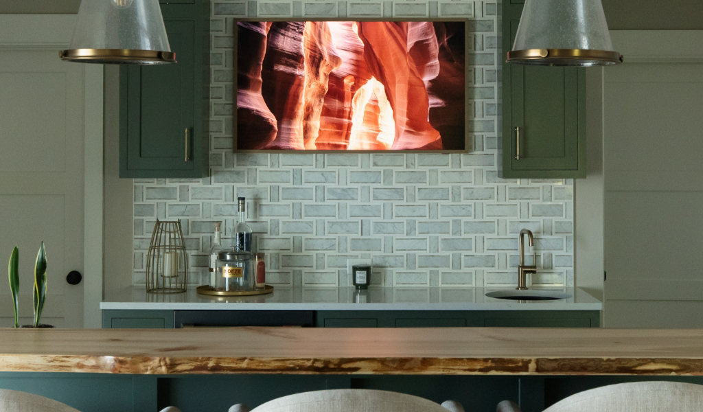 Satkoski – 47 A modern kitchen from a Byron Center Whole Home Remodel features green cabinets, a wooden counter, tile backsplash, a mounted TV displaying abstract art, and metal pendant lights.