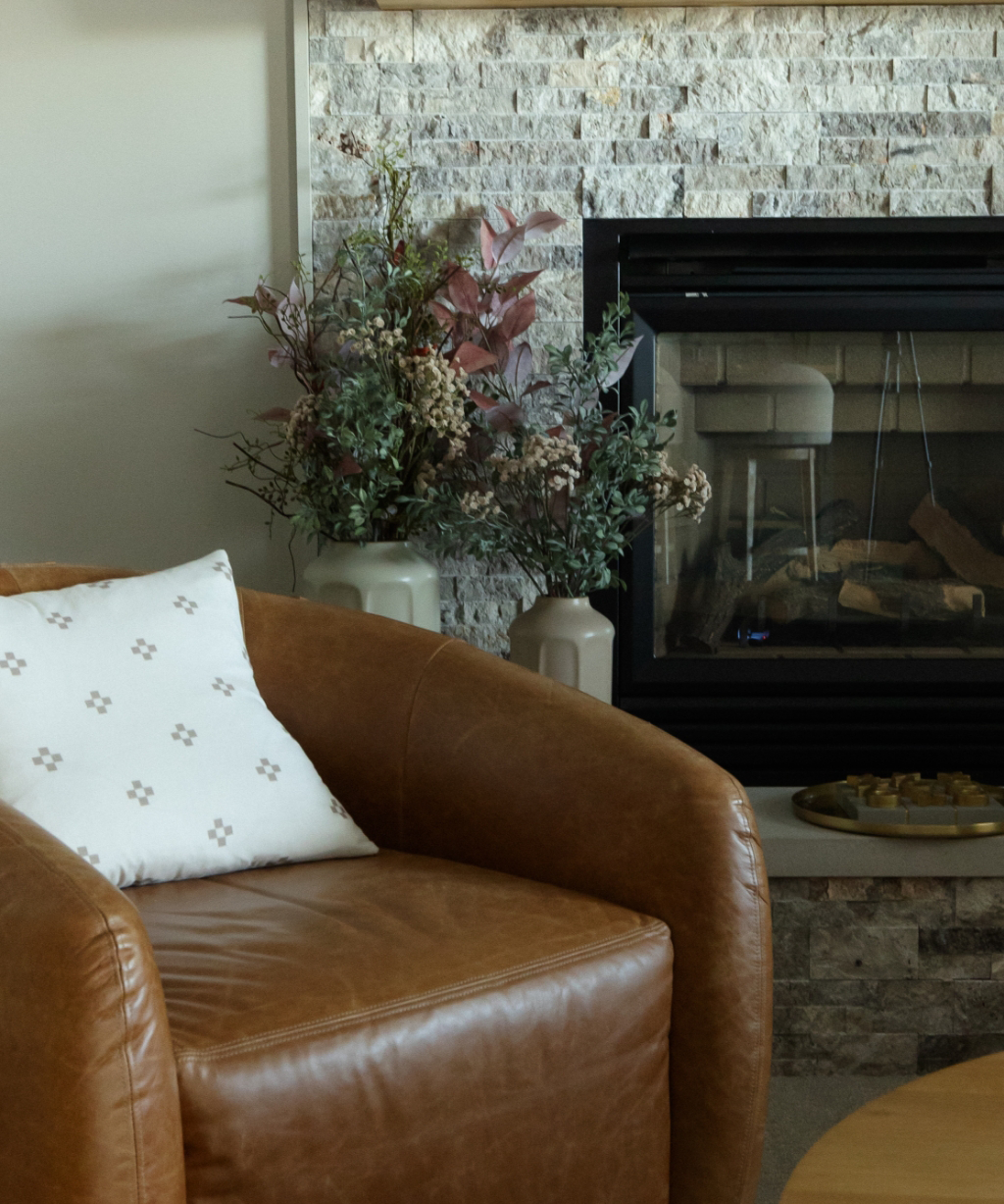 A brown leather armchair with a white patterned pillow sits next to a stone fireplace, styled for a Byron Center Whole Home Remodel, with vases of dried flowers placed on the hearth.