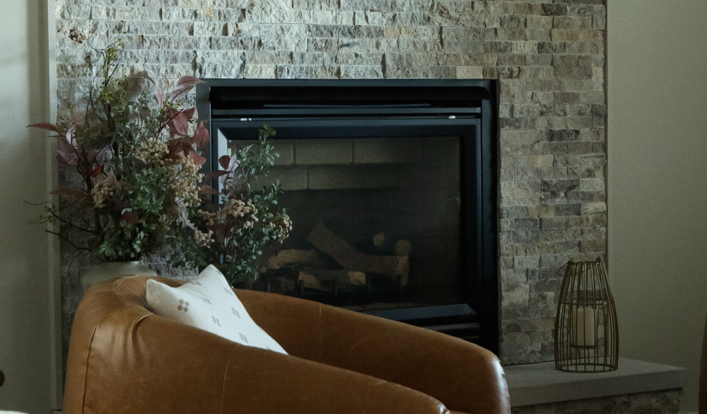 A brown leather chair sits in front of a stone fireplace, featuring a floral arrangement and decorative lantern on the hearth—an inviting space inspired by a recent Byron Center Whole Home Remodel.