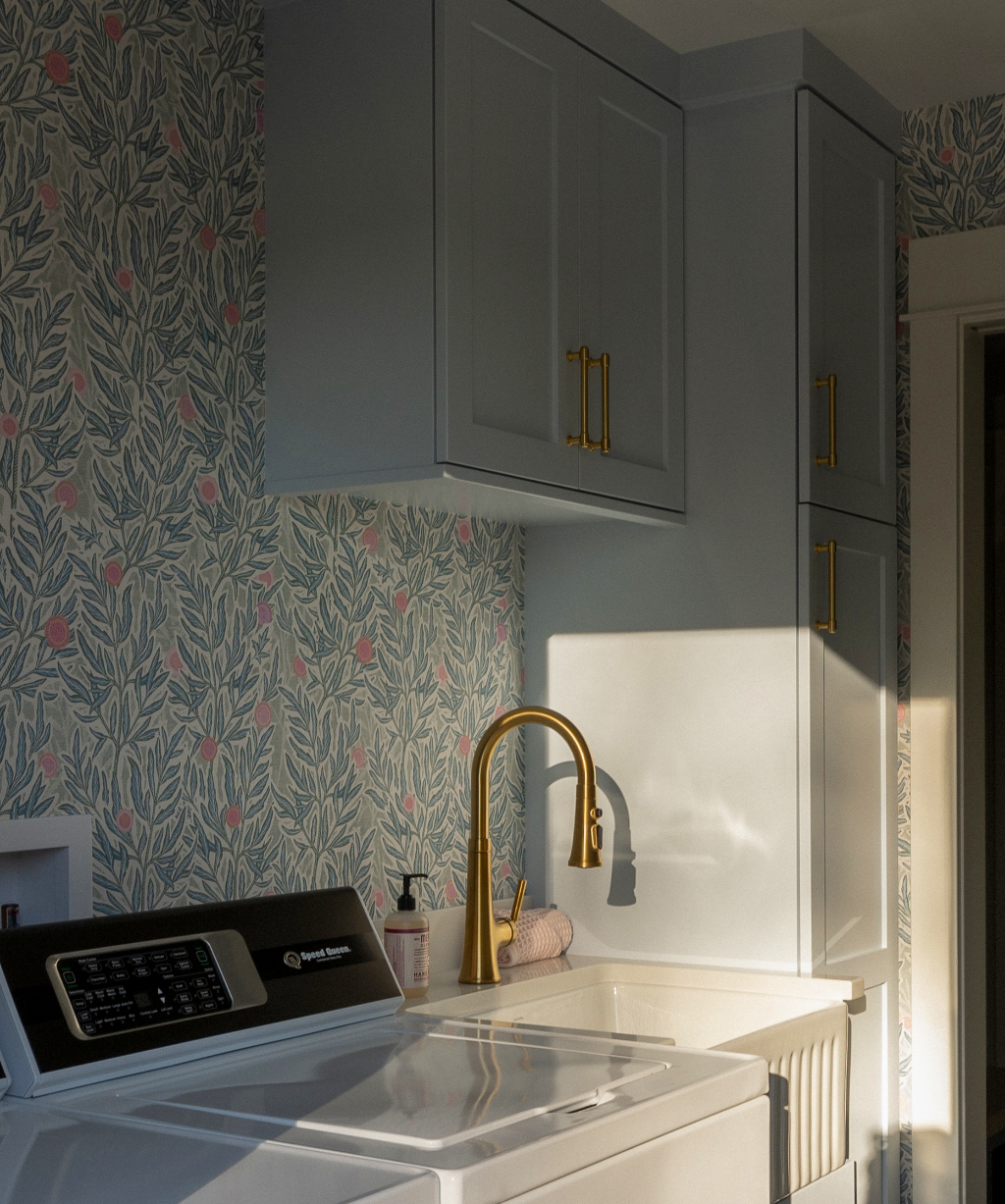 Satkoski – 5 A laundry room from a Byron Center Whole Home Remodel features a washing machine, dryer, gold faucet, blue cabinets with gold handles, and floral wallpaper. Sunlight casts beautiful shadows on the appliances and wall.