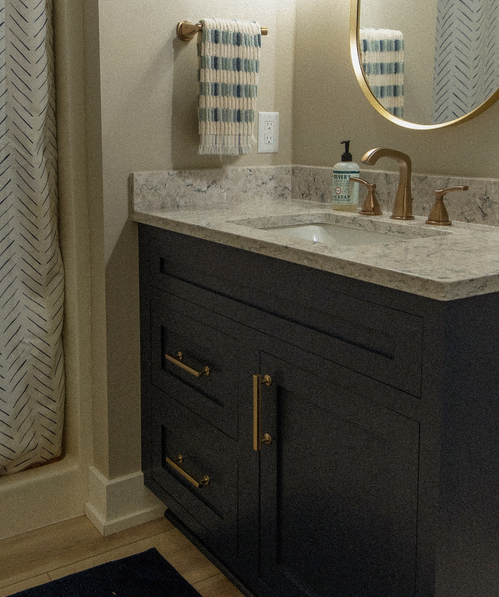 A bathroom from a Byron Center Whole Home Remodel features a dark vanity with gold handles, a marble countertop, a round mirror, a gold faucet, a striped towel, and a blue and white shower curtain.