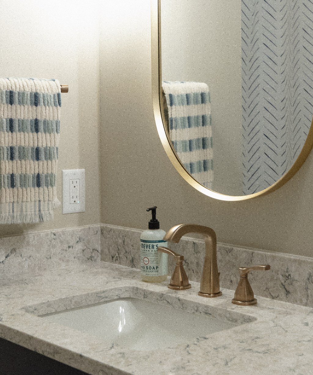 Bathroom sink with a gold faucet, marble countertop, soap dispenser, striped towel on a rack, and a round mirror above—part of a stunning Byron Center Whole Home Remodel.
