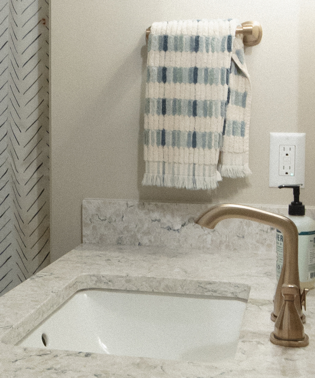 Beige bathroom countertop with a built-in sink, gold faucet, soap dispenser, towel on a rack, and patterned shower curtain in the background—part of a stylish Byron Center Whole Home Remodel.