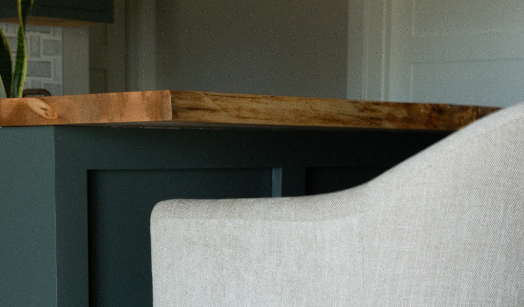 A beige upholstered chair sits in front of a dark green kitchen island with a wooden countertop, showcasing the elegance of a Byron Center Whole Home Remodel.