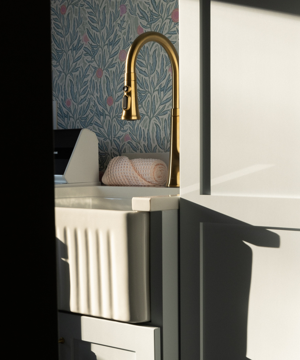 Satkoski – 6 A white farmhouse sink with a gold faucet is set against floral wallpaper in this Byron Center Whole Home Remodel. A rolled towel sits on the countertop beside the sink, as sunlight casts shadows on the cabinetry.