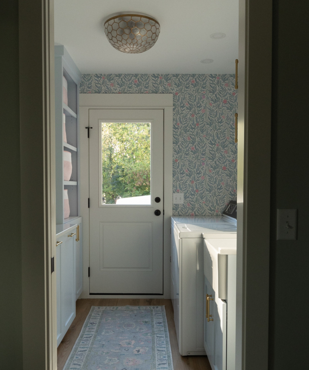 A laundry room from a Byron Center Whole Home Remodel features patterned wallpaper, white cabinets, a stylish light fixture, a cozy rug, and a door with a window letting in natural light.
