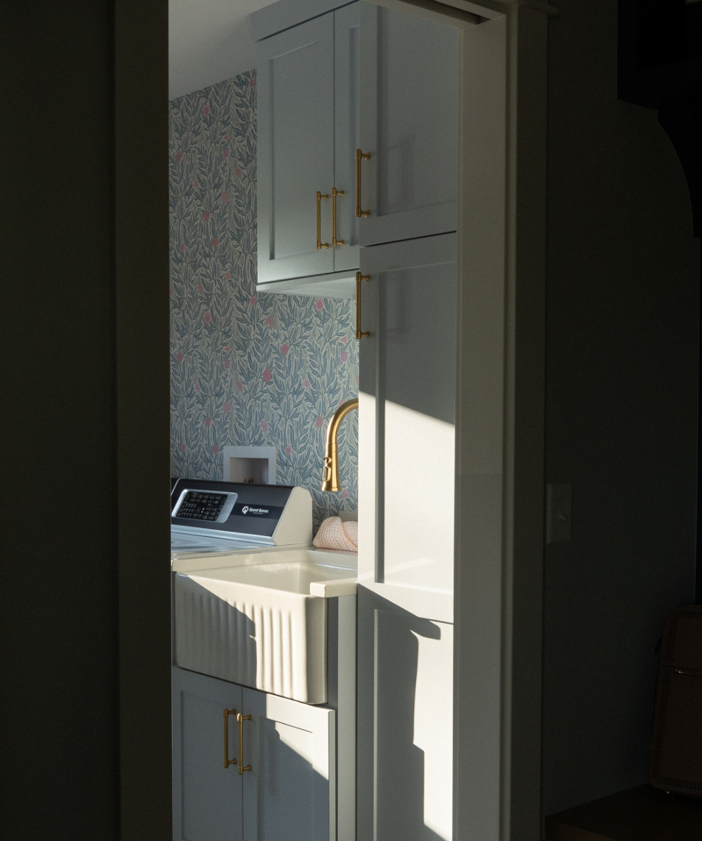 Satkoski – 8 Sunlight streams into this Byron Center Whole Home Remodel laundry room, featuring light gray cabinets, a farmhouse sink with a brass faucet, and a washing machine set against floral-patterned wallpaper.