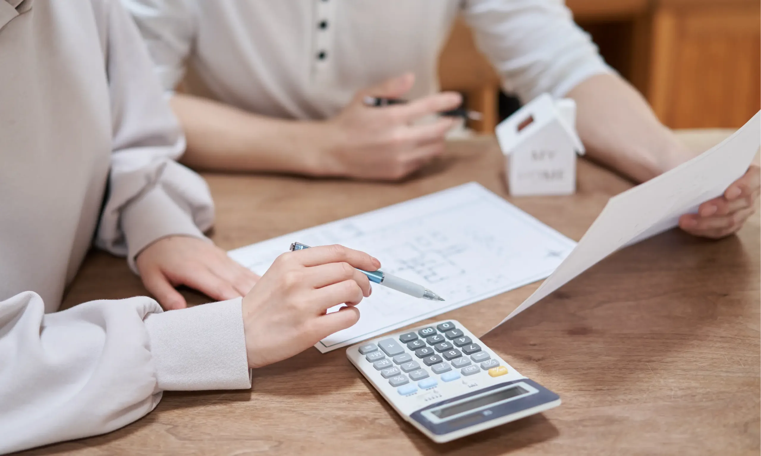 Two people review documents at a table with a calculator and a small house model, suggesting financial planning or home buying—perhaps even budgeting for the latest kitchen design trends.