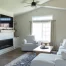Bright, modern living room with vaulted ceilings, neutral furniture, a wall-mounted TV above a sleek fireplace, floating wood shelves, and large windows letting in natural light.