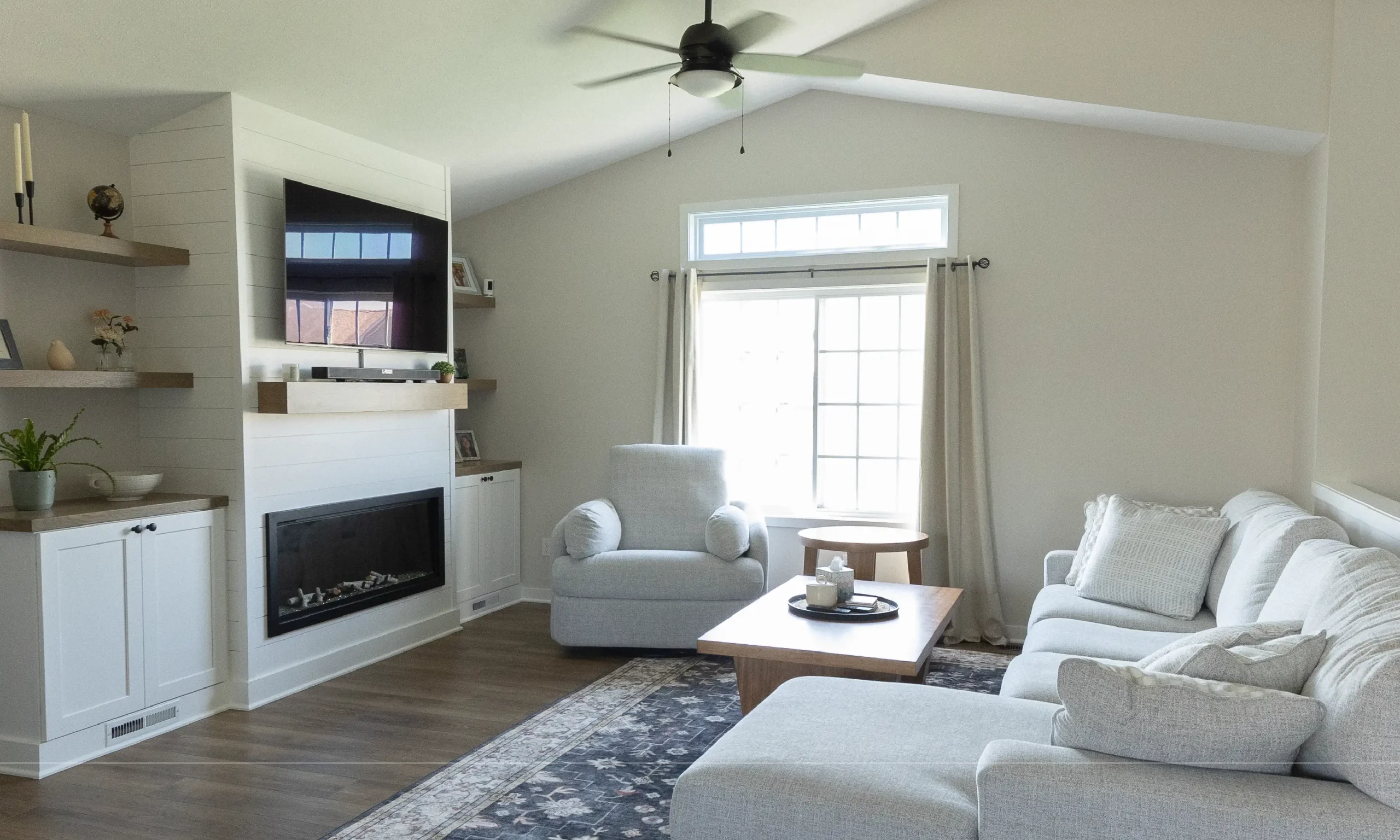 Bright, modern living room with vaulted ceilings, neutral furniture, a wall-mounted TV above a sleek fireplace, floating wood shelves, and large windows letting in natural light.